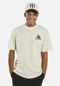 Beige cotton t-shirt featuring a black triangle graphic with an eye design and text "REDEFINED REBEL." Model wears it with hands in pockets.