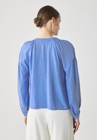 Lys blå, stripete bluse med lange ermer, samlet detaljer på skuldrene og elastiske mansjetter. Glatt tekstur, litt løs passform.