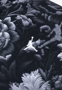 Czarny materiał z bogatym wzorem kwiatowym w odcieniach szarości, z białym logo Jordana wyraźnie widocznym wśród wzoru.