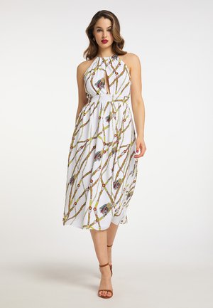 Donna che indossa un vestito midi bianco con collo halter con stampa colorata a catene e nappine, abbinato a tacchi marroni con cinturino alla caviglia, in piedi su sfondo bianco.