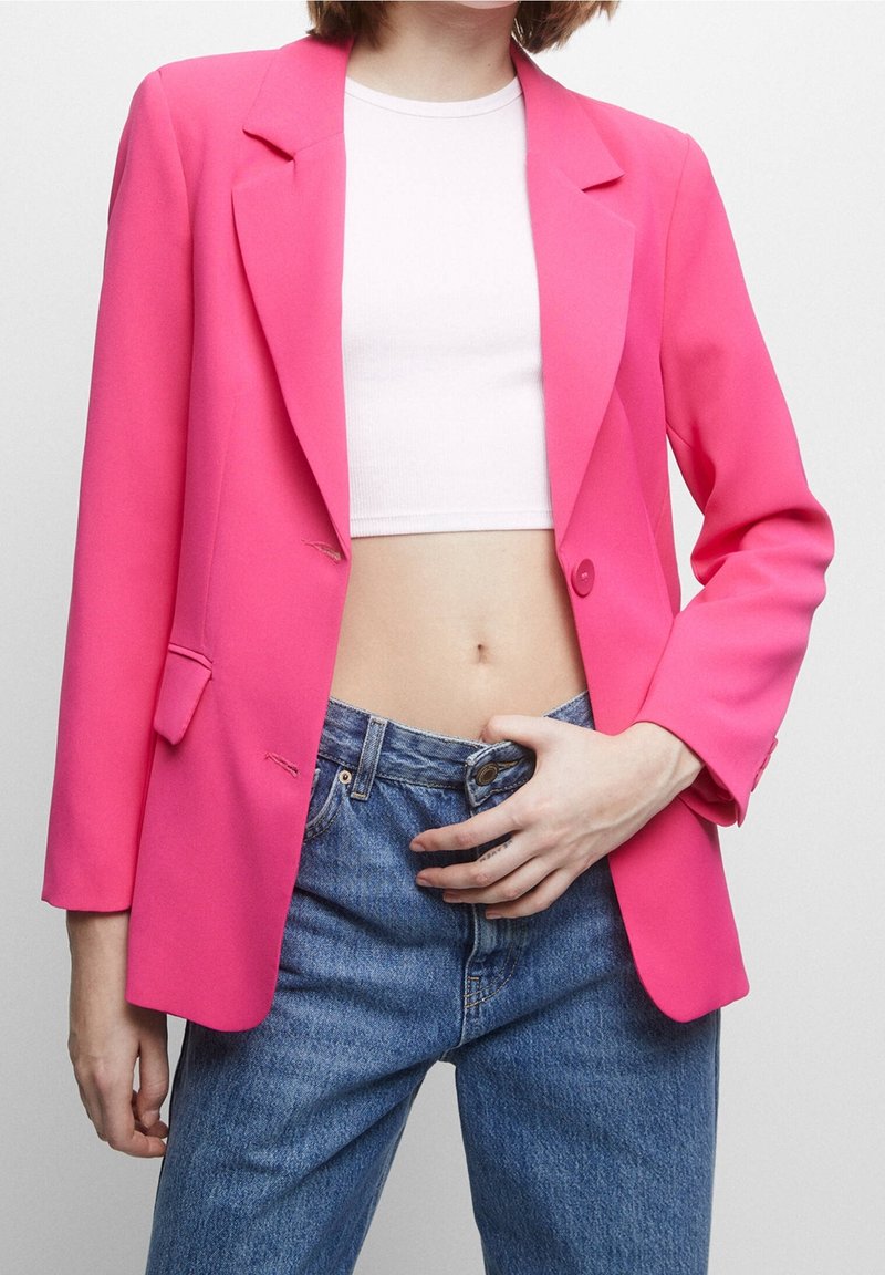 Femme portant un blazer rose vif sur un crop top blanc, en train de boutonner un jean bleu taille haute, sur fond clair uni.