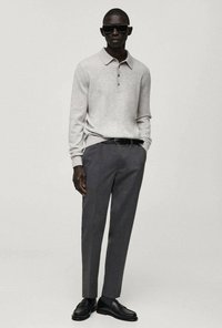 Helles graues Polohemd mit Knöpfen, kombiniert mit dunkelgrauen Hosen und schwarzen Slippern. Das Modell trägt Sonnenbrillen und steht mit den Händen in den Taschen.