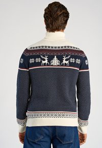Marinblå stickad tröja med vitt Fair Isle-mönster, med renar och snöflingor, hög krage och ribbad nederkant med röda och beiga detaljer.