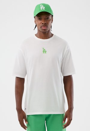 Uomo che indossa un cappellino da baseball verde con il logo LA, una T-shirt bianca con il logo verde LA e pantaloncini verdi, in piedi davanti a uno sfondo bianco uniforme.