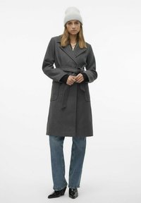 Cappotto in misto lana grigia con cintura, colletto a revers e tasche laterali. Abbinato a jeans blu e stivali neri con tacco, completato da un berretto grigio chiaro.