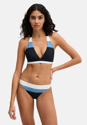 Frau, die einen schwarz-blau-weißen gestreiften Neckholder-Bikini trägt, mit der Hand in der Hüfte stehend vor einfachem hellem Hintergrund.