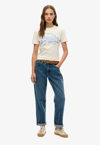 T-shirt en coton couleur crème avec logo SuperDry bleu, associé à un jean taille haute bleu et des baskets beiges. Les poignets du jean sont retroussés.