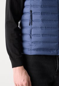 Chaleco acolchado azul con costuras horizontales, que presenta un bolsillo lateral con cremallera y una textura suave, llevado sobre una camiseta de manga larga negra.