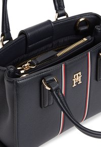 Μαύρη τσάντα χειρός από δερματίνη με υφή, με χρυστό φερμουάρ, διπλές λαβές και κάθετη λεπτομέρεια με κόκκινη-λευκή-μαύρη ρίγα στο μπροστινό πάνελ.