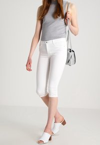 Jean blanc coupé avec une coupe slim, associé à un top sans manches gris. Le modèle porte des mules blanches à bout ouvert et tient un sac gris.