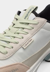 Κοντινό πλάνο μπεζ και ανοιχτοπράσινου αθλητικού παπουτσιού Calvin Klein με υφασμάτινη ετικέτα, επενδυμένο κολάρο και υφασμένα σουέτ διακοσμητικά σε λευκή σόλα.