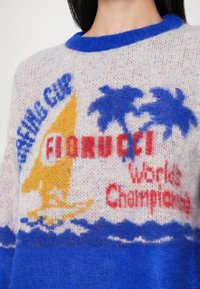 Femme portant un pull douillet avec un voilier, deux palmiers et les inscriptions "RACING CLUB", "FIORUCCI" et "World Champions."