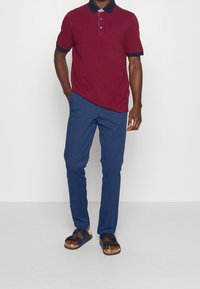 Burgundy pikétröja med marinblå krage, kort ärm, kombinerad med blå byxor och bruna sandaler med dubbel remdesign.