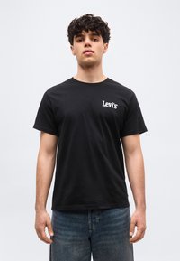 Νεαρός άντρας με σγουρά μαλλιά, φορώντας μαύρο t-shirt Levi's και μπλε τζιν, στέκεται μπροστά σε απλό ανοιχτό γκρι φόντο.