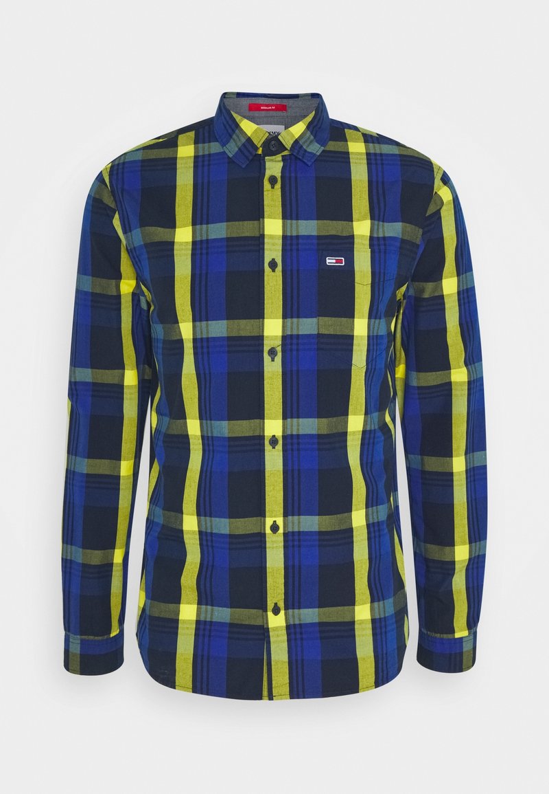 Chemise à manches longues avec boutons, motif à carreaux bleu, jaune et noir et petit écusson avec logo sur la poitrine, présentée sur fond blanc.