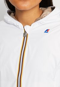 Personne portant une veste blanche avec une fermeture éclair à rayures jaune, bleu marine et blanche et un petit logo rouge et bleu sur la poitrine près de l'épaule.