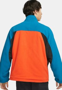 Fleecejacka med blå krage, orange rygg och svarta sidoflikar. Mjuk textur med avslappnad passform och inga synliga märken.