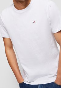 Vit bomullst-shirt med korta ärmar, rund halsringning och en liten logotyp på bröstet med röda, vita och marinblå ränder.