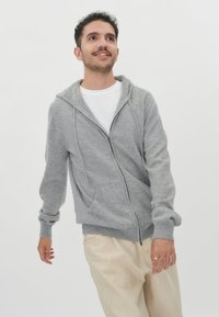 Grå zip-up hoodie med snörning i huvan, ribbade avslut och en framlapp, tillsammans med ljusa byxor. Slät textur.