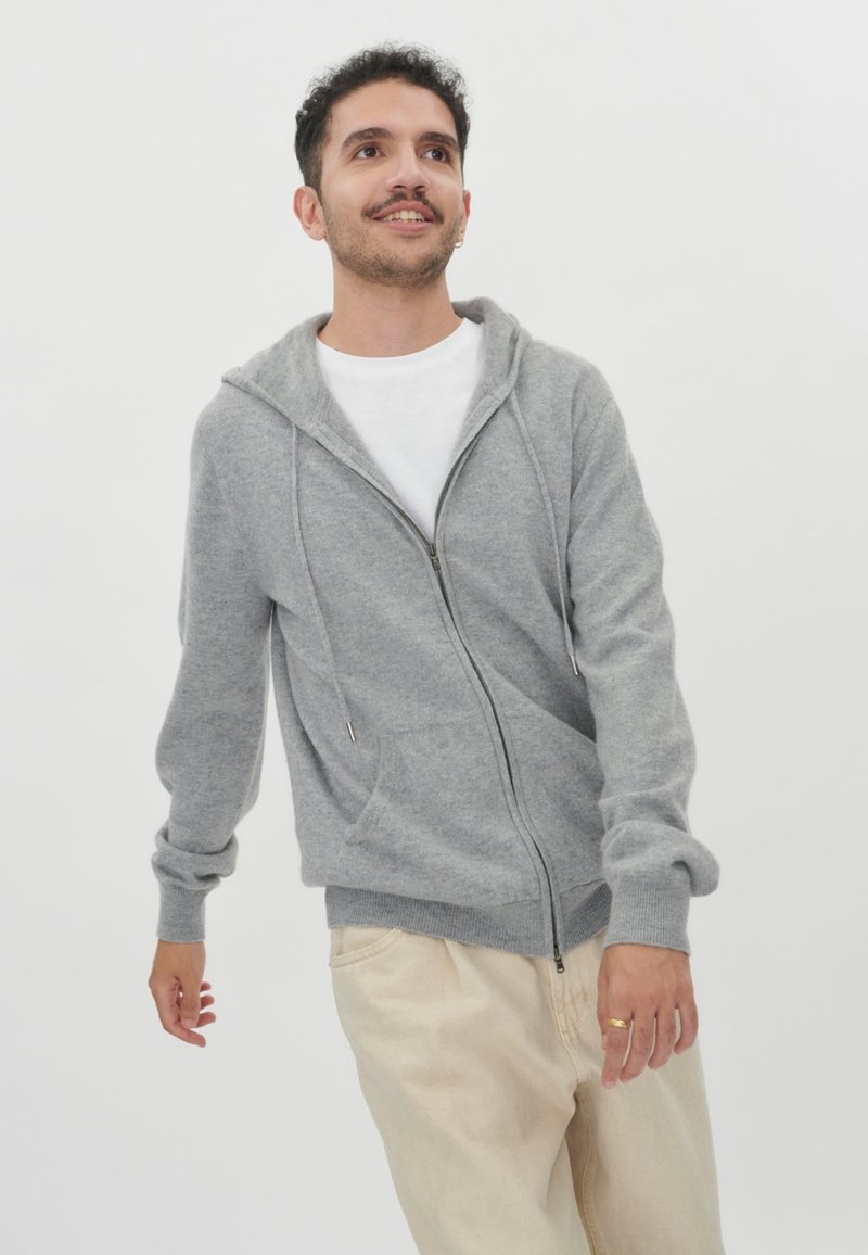 Grå zip-up hoodie med snörning i huvan, ribbade avslut och en framlapp, tillsammans med ljusa byxor. Slät textur.