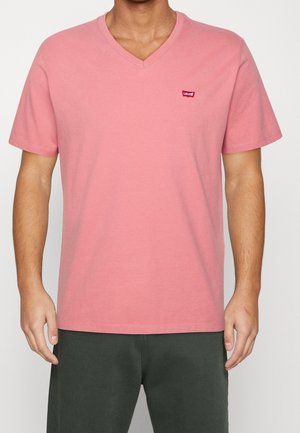 Homme portant un T-shirt Levi’s rose col V à manches courtes et un pantalon vert foncé, debout devant un fond uni.