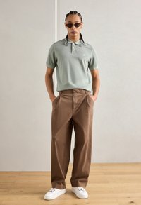 Lys grønn polo skjorte med korte ermer og knapper, kombinert med brune vide bukser og hvite sneakers. Glatt stoff, minimal merking.