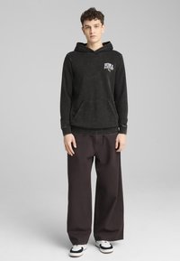 Felpa nera con tasca a marsupio e logo bianco, abbinata a pantaloni larghi scuri e scarpe da ginnastica con dettagli in bianco e nero.