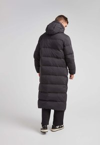 Lange zwarte pufferjas met een capuchon, met horizontale gestikte secties, zijzakken met ritssluiting en een rechte snit.