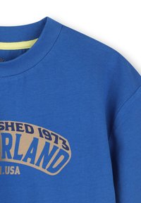 Blauwe katoenen t-shirt met ronde hals en korte mouwen. Voorzien van een gouden grafisch ontwerp met de tekst "Opgericht 1973 Timberland USA" op de voorkant.
