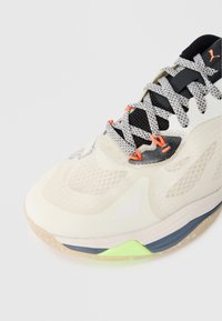 Fény bézs sportcipő texturált hálós felsőrésszel, fekete részletekkel, narancssárga fűzőlyukakkal és zöld sarokdíszítéssel. Átlátszó gumitalp.