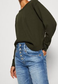 Haut en ribbed kaki à manches longues associé à un jean taille haute bleu clair avec un devant à boutons asymétriques et des accents déchirés.