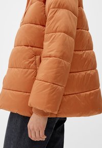 Osoba w pomarańczowej, pikowanej kurtce puchowej nosząca ciemne dżinsy, z widoczną ręką swobodnie opartą przy boku na białym tle.