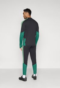 Μαύρη αθλητική φόρμα με πράσινες λεπτομέρειες, μακριά μανίκια με υφή υφάσματος και τρεις πράσινες ρίγες στα πλάγια του παντελονιού.