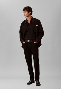 Νέος άνδρας στέκεται με τα χέρια στις τσέπες, φοράει μαύρο μπουφάν, μαύρο πουκάμισο, μαύρο παντελόνι, μαύρη ζώνη και μαύρα παπούτσια, μπροστά σε γκρίζο φόντο.