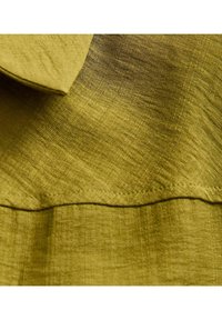 Tessuto verde oliva con finitura strutturata. Presenta un colletto piegato e una cucitura visibile lungo l'orlo inferiore.