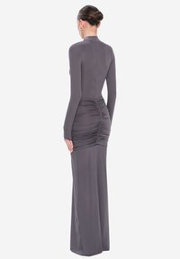 Mujer con el cabello recogido en un moño, vistiendo un vestido gris de manga larga ajustado, con detalles fruncidos en la parte posterior y un dobladillo hasta el suelo.