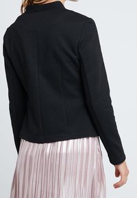 Czarna, dopasowana marynarka z wysokim kołnierzem, o gładkiej fakturze i wyraźnym kroju, zestawiona z różową, plisowaną spódnicą.