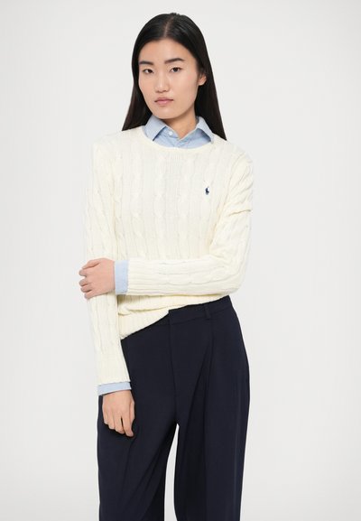 Kremni pulover z vzorcem kaveljčnih pletenic čez svetlo modro srajco z ovratnikom, kombiniran s temno modrimi hlačami. Opazne teksture in majhen logotip na puloverju.