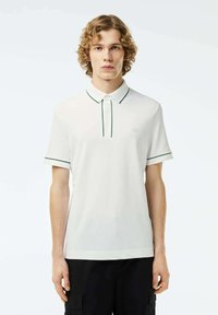 Vit pikétröja med krage och knäppning samt gröna detaljer runt kragen och ärmarna. Mjuk och slät tygtextur.