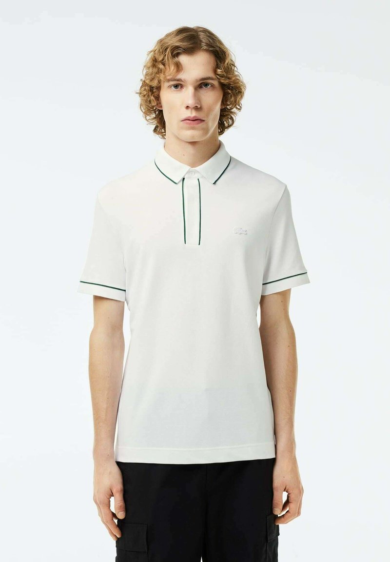 Vit pikétröja med krage och knäppning samt gröna detaljer runt kragen och ärmarna. Mjuk och slät tygtextur.