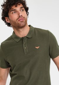 Polo shirt verde oliva in cotone, con colletto, tre bottoni e un piccolo logo ricamato color corallo sul petto.