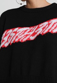 Schwarzer Strickpullover mit einem strukturierten roten und weißen Design über der Brust, mit rundem Halsausschnitt und gerippten Bündchen.
