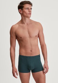 Boxer da uomo verde scuro con una texture liscia, cintura elastica e cuciture minime, indossati da un modello su uno sfondo neutro.