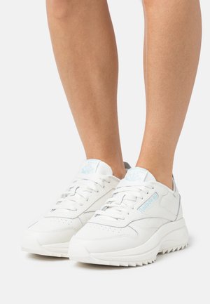Personne portant des baskets Reebok Classic blanches avec un logo bleu clair, se tenant sur un fond blanc.