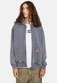 Grå hættebluse med frontlommer, der har et tekstureret stof og et logo-patch på højre side. Bæres over en hvid t-shirt.