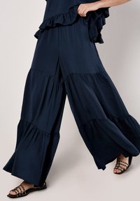 Navy blue wide-leg pants with ruffle accents, featuring a tiered design and lightweight fabric. Strappy black sandals visible at the bottom.