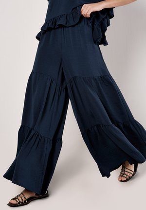 Navy blue wide-leg pants with ruffle accents, featuring a tiered design and lightweight fabric. Strappy black sandals visible at the bottom.