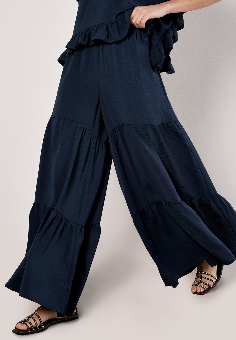 Navy blue wide-leg pants with ruffle accents, featuring a tiered design and lightweight fabric. Strappy black sandals visible at the bottom.
