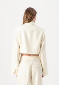 Blazer corto de crema con un patrón floral texturizado, hombros estructurados y mangas largas, combinado con pantalones de talle alto a juego.