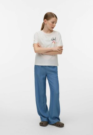 Jeune personne aux cheveux châtain clair attachés en queue de cheval, portant un t-shirt blanc avec un motif graphique, un pantalon bleu ample et des chaussures type sabots marron, debout les bras croisés.